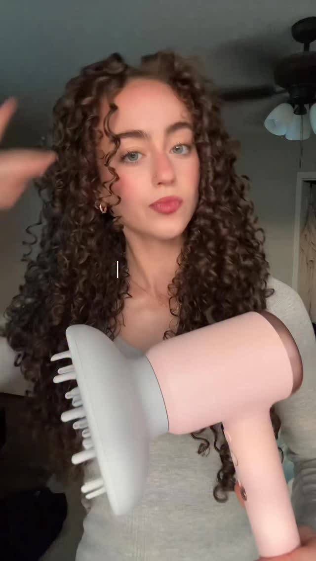 Woman with curly brown hair holding a pink hair dryer with a white diffuser.