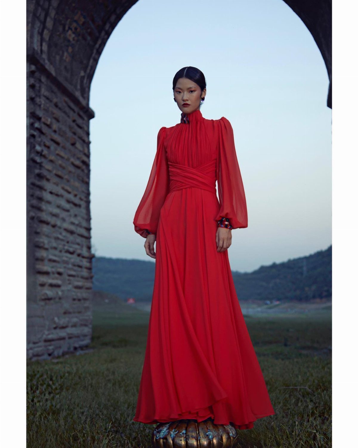Woman in a red long-sleeved gown with draped bodice under a stone archway