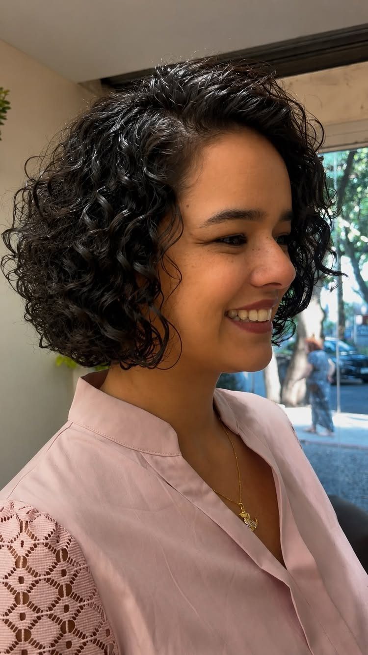 18 Medium Length Curly Hair Hairstyles, From Bobs to Shags 8 Woman with a classic curly bob hairstyle smiling in a pink blouse.