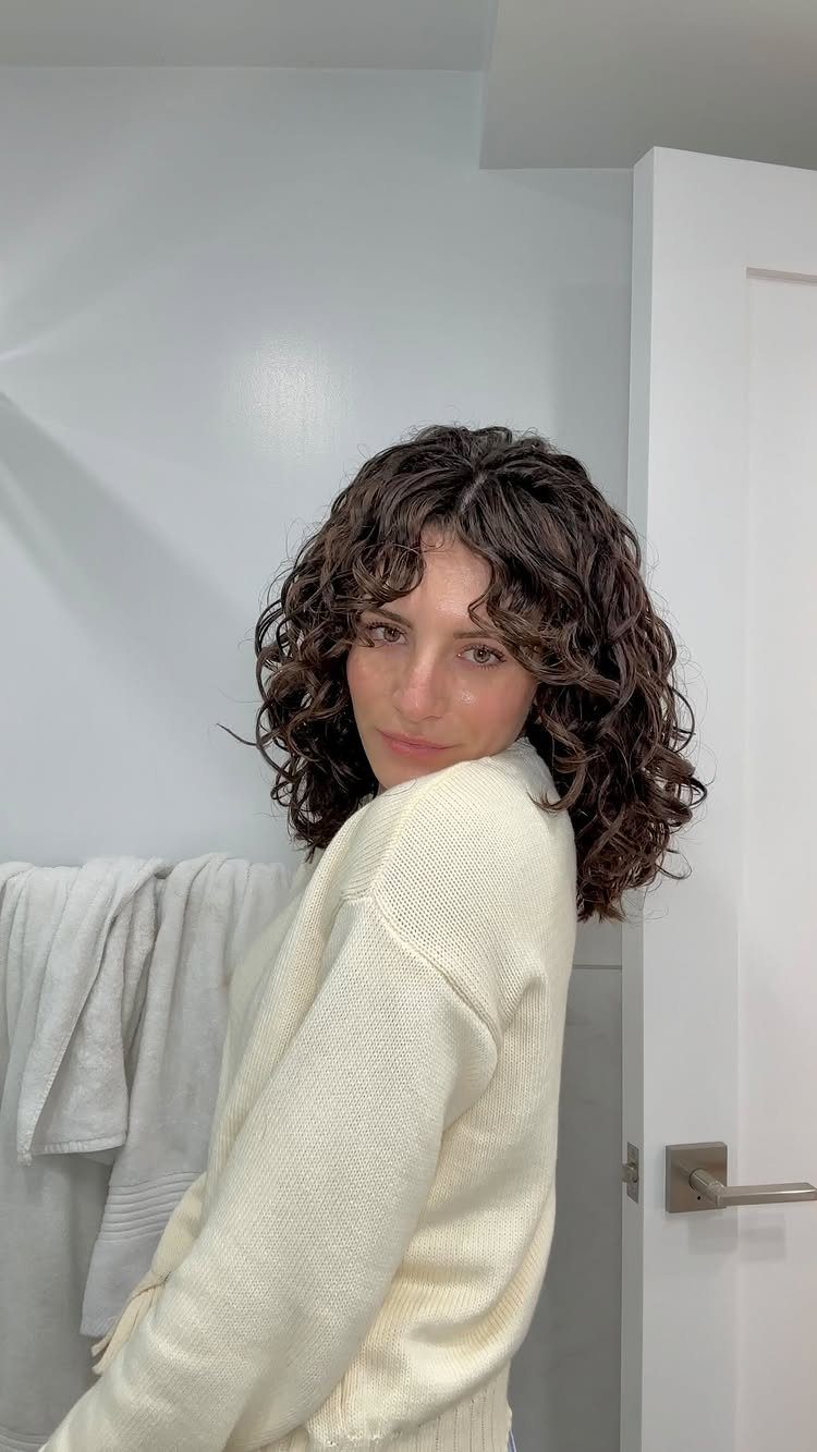 18 Medium Length Curly Hair Hairstyles, From Bobs to Shags 7 Woman with medium length curly brown hair and curtain bangs wearing cream sweater.