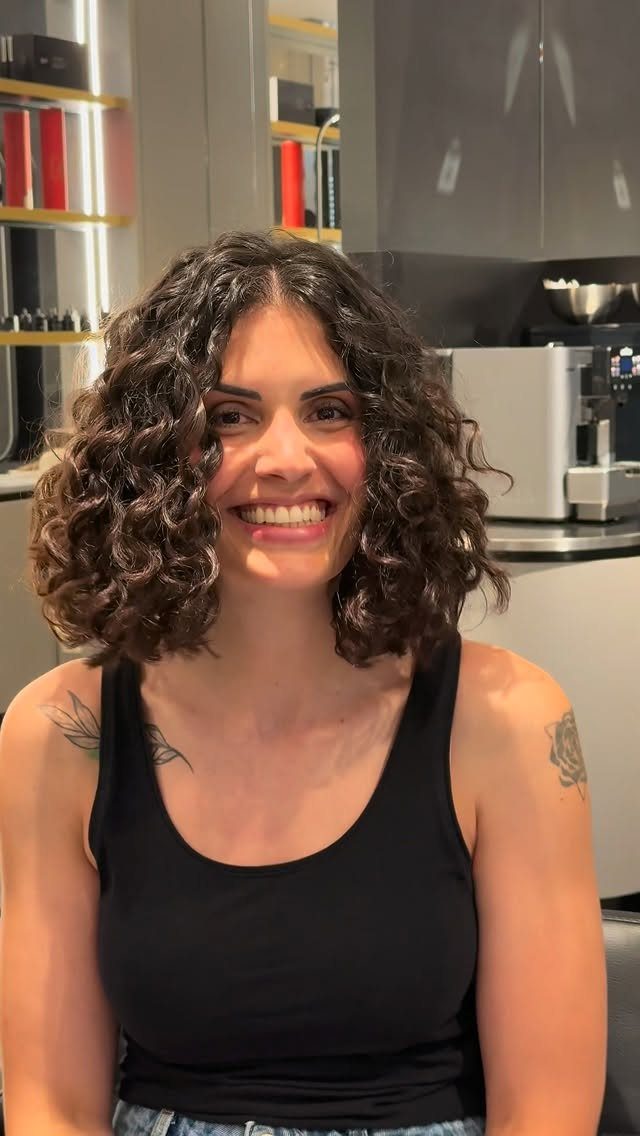 18 Medium Length Curly Hair Hairstyles, From Bobs to Shags 4 Woman with medium length dark curly hair smiling in a modern kitchen setting.