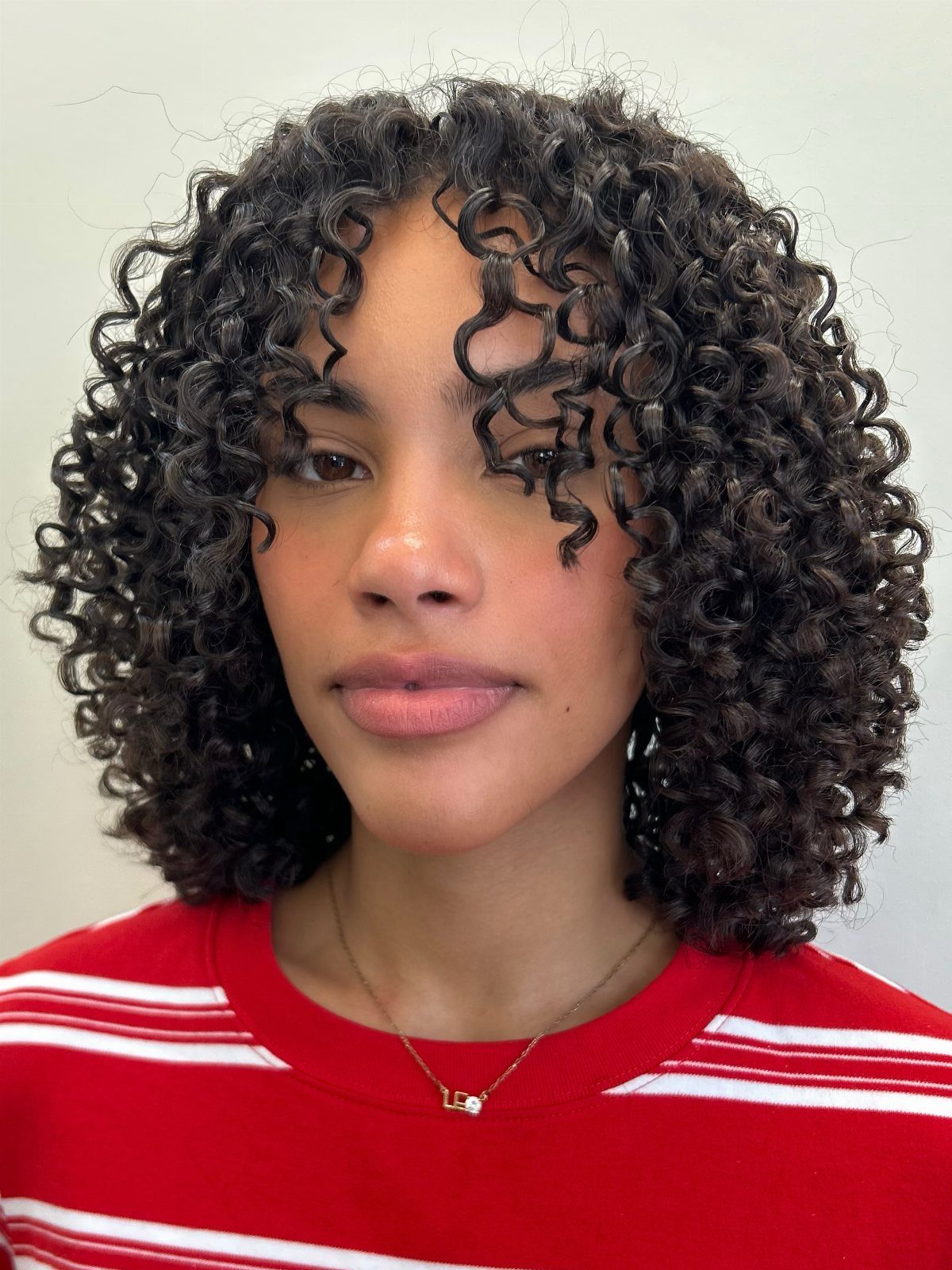 18 Medium Length Curly Hair Hairstyles, From Bobs to Shags 14 Woman with tight dark ringlets and curly bangs wearing a red striped shirt.