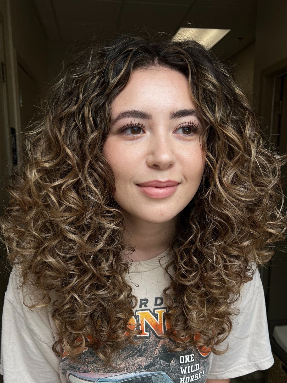 18 Medium Length Curly Hair Hairstyles, From Bobs to Shags 12 Woman with shoulder-length curly brown hair and golden-blonde balayage highlights posing indoors.