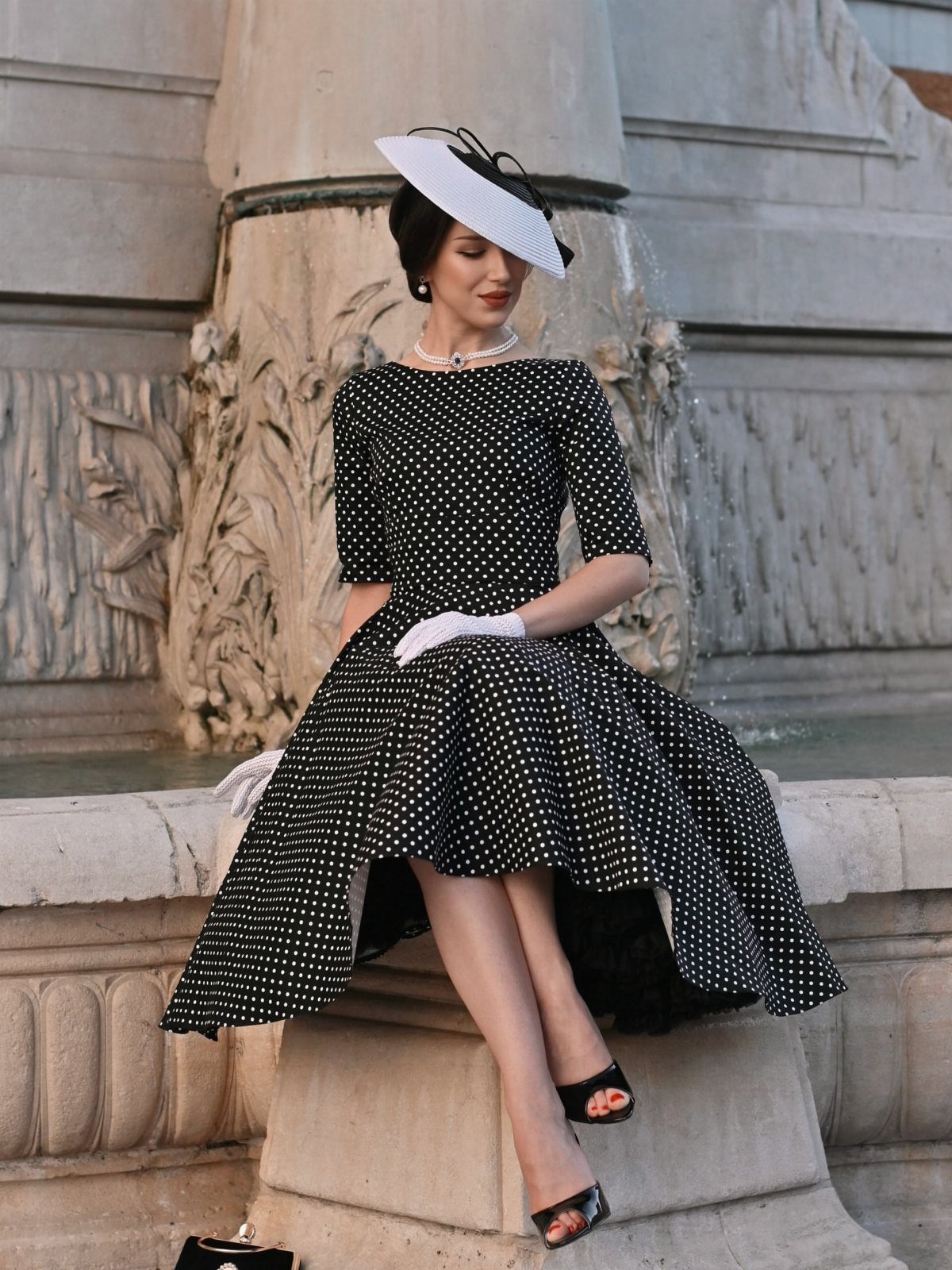 Woman in a black and white polka dot vintage dress sitting on stone fountain.