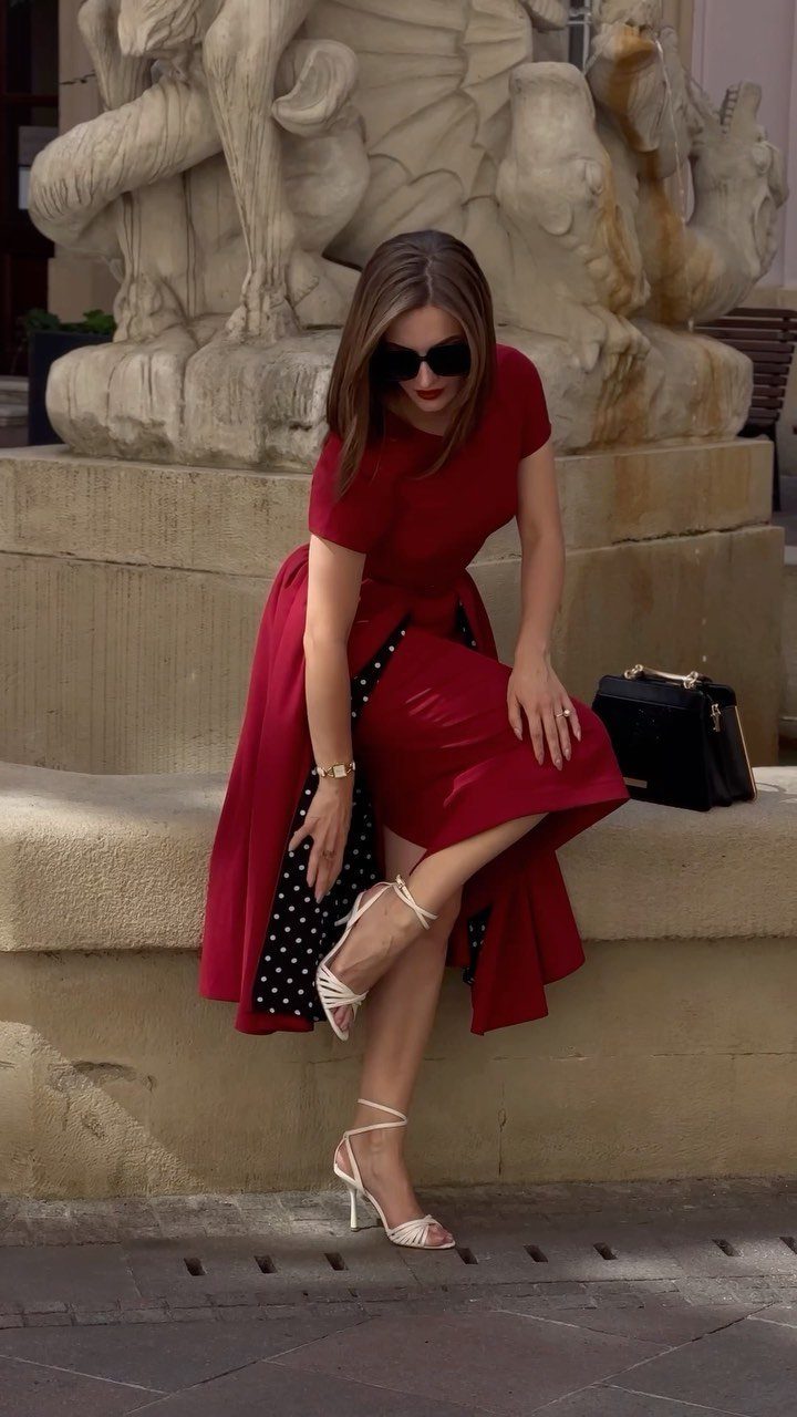 Woman in a short red dress with a long overskirt sitting by a sculpture.