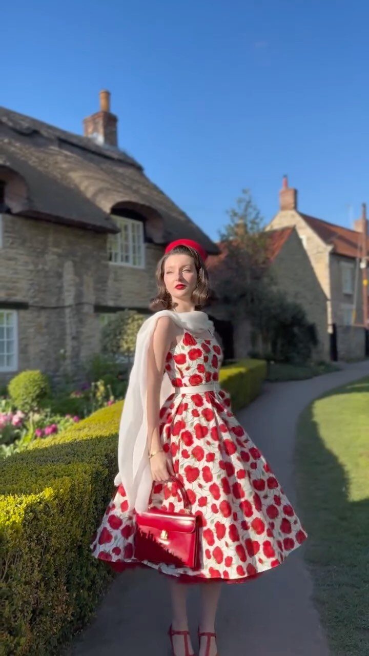 Woman in a red heart-patterned short vintage dress standing by a thatched cottage.