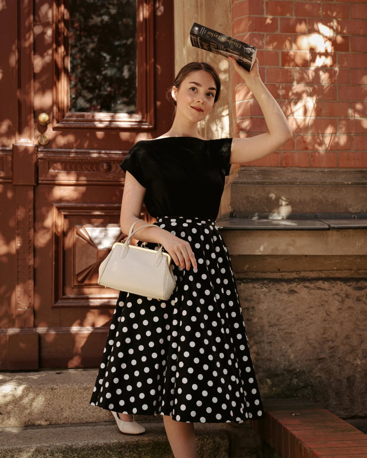Woman in a black wide-neckline dress with a full white polka-dot skirt holding a book.