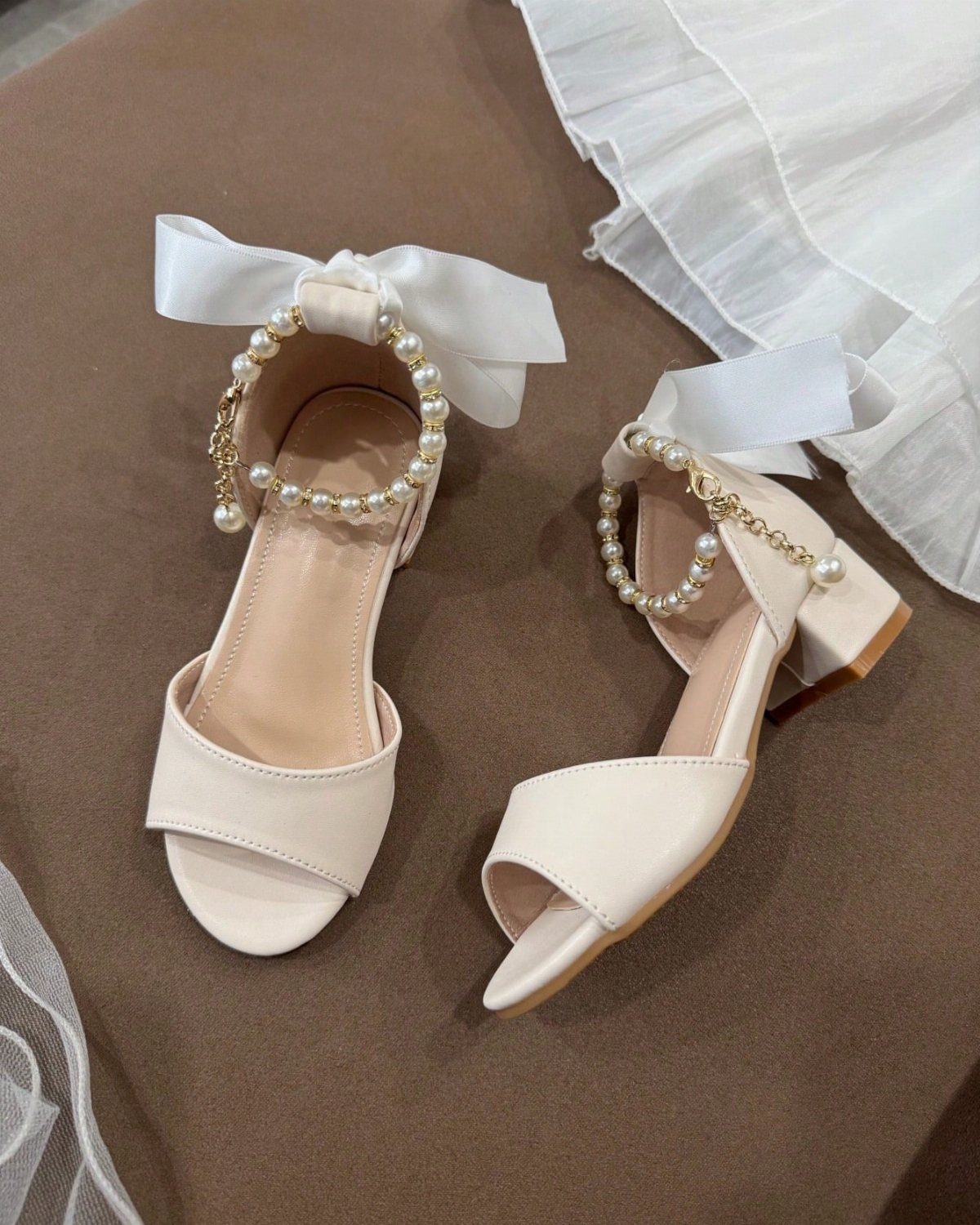 Cream heeled sandals with pearl ankle straps and white bows on brown surface