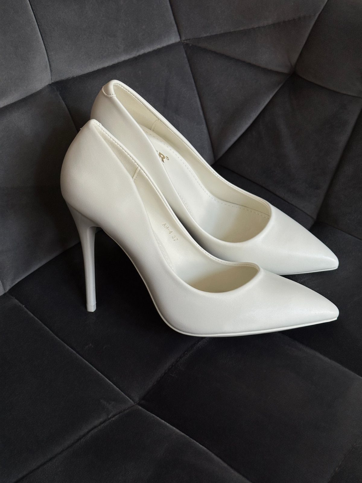 White pointed-toe high-heeled pumps resting on a dark grey quilted velvet background.