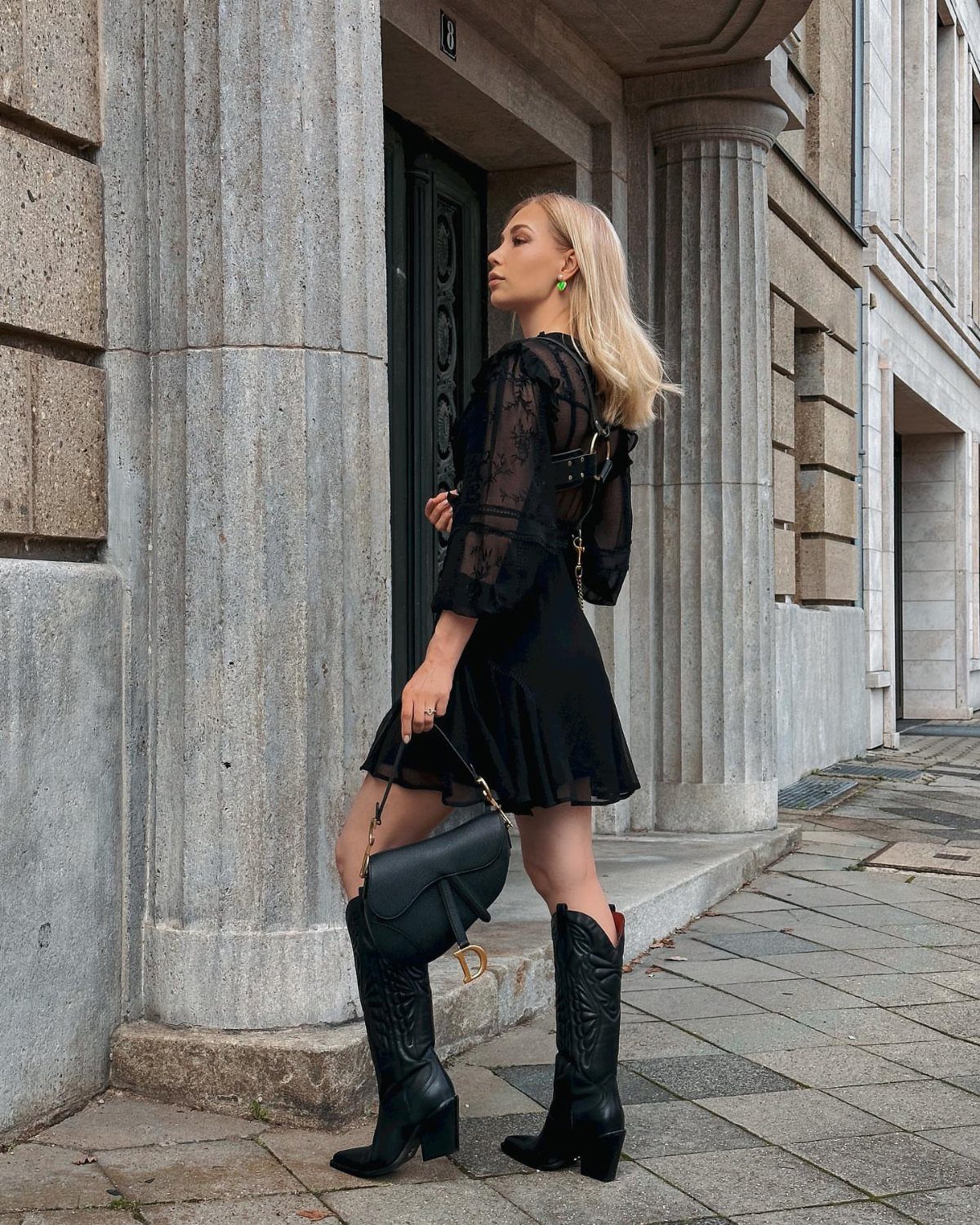 29 Rodeo Inspired Outfits Built on Fringe, Denim, and Boots 26 Woman in a short black dress and black cowboy boots posing by stone columns.
