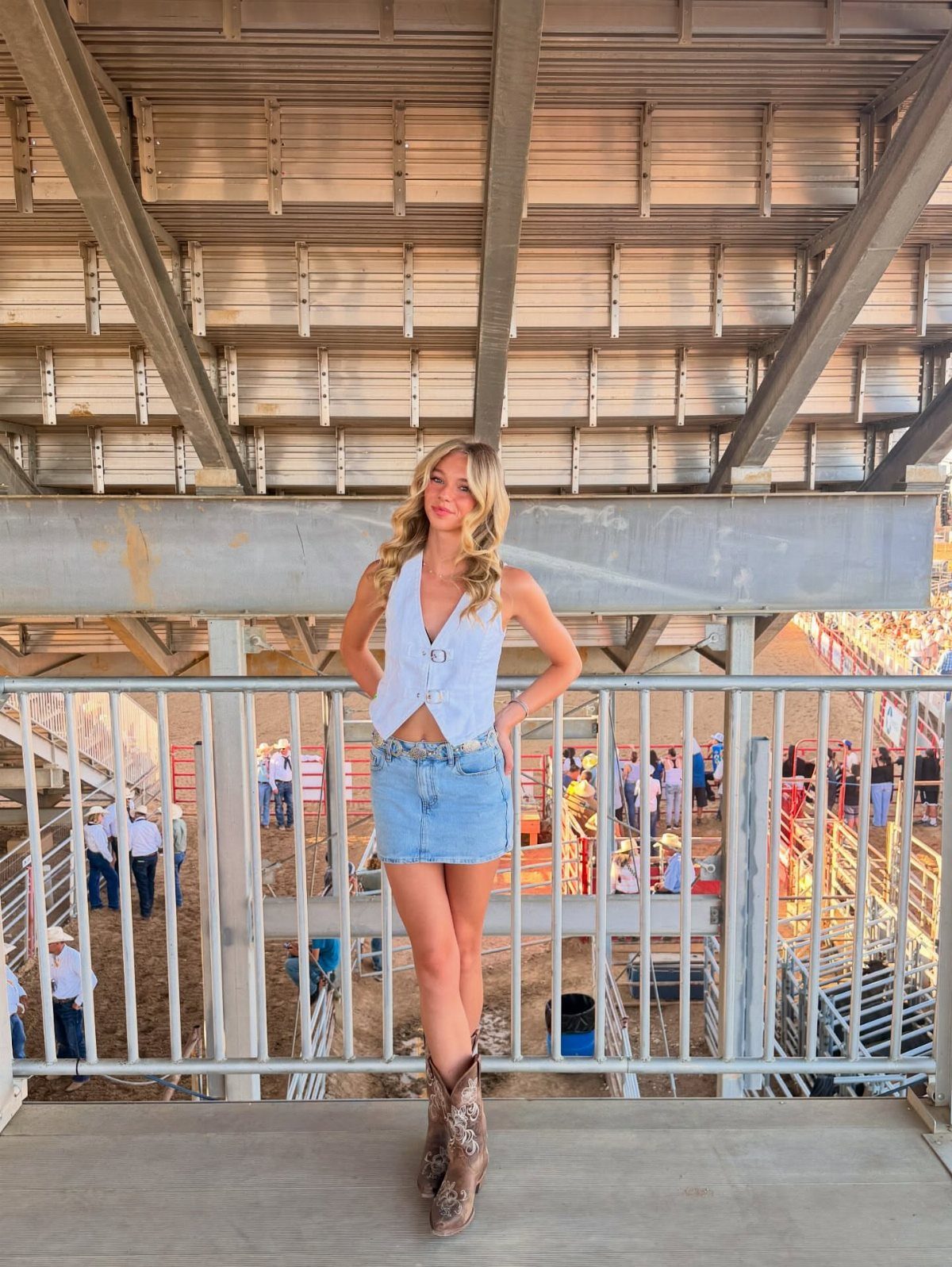 29 Rodeo Inspired Outfits Built on Fringe, Denim, and Boots 5 Woman wearing a white sleeveless shirt, denim mini skirt, and brown cowboy boots.