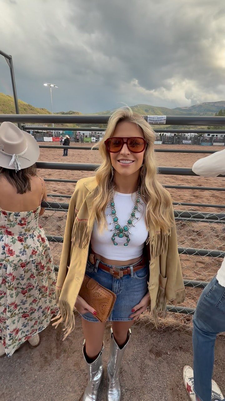29 Rodeo Inspired Outfits Built on Fringe, Denim, and Boots 3 Woman in tan fringe jacket, denim skirt, and silver cowboy boots at a rodeo.