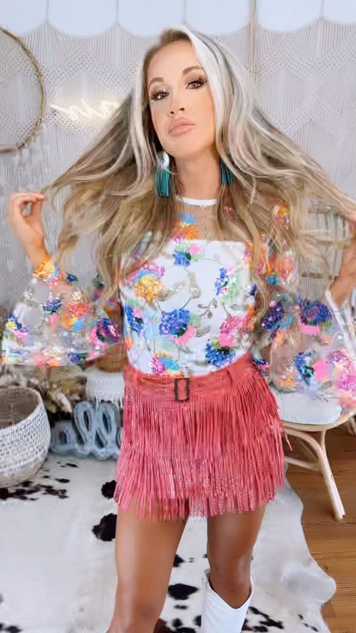 29 Rodeo Inspired Outfits Built on Fringe, Denim, and Boots 9 Woman wearing a pink fringed mini skirt, floral blouse, and white cowboy boots.