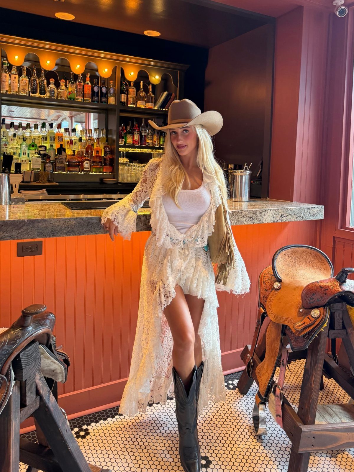 29 Rodeo Inspired Outfits Built on Fringe, Denim, and Boots 16 Woman in white lace dress and tan cowboy hat posing in Western bar.