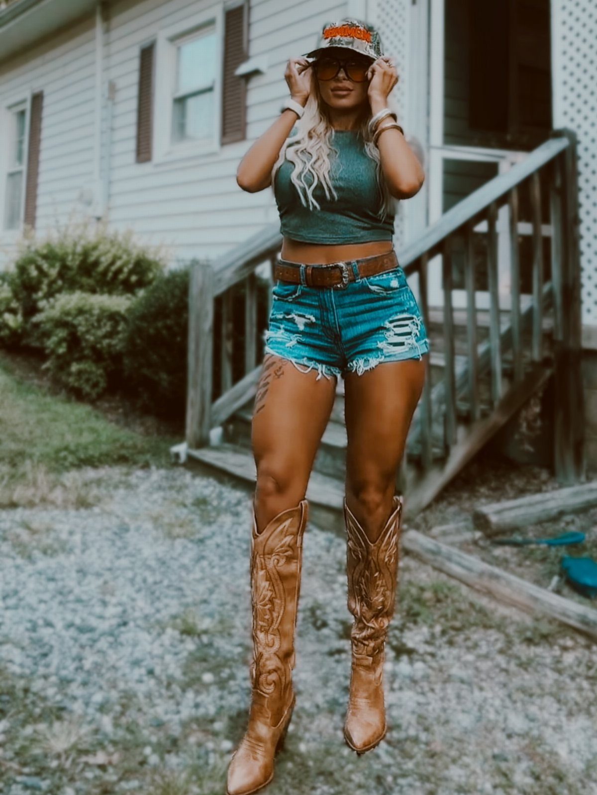 29 Rodeo Inspired Outfits Built on Fringe, Denim, and Boots 27 Woman in green tank top, denim shorts, and tall brown cowboy boots posing outdoors.