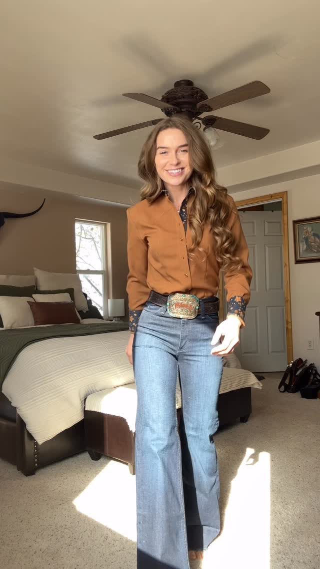 29 Rodeo Inspired Outfits Built on Fringe, Denim, and Boots 13 Woman wearing a brown Western shirt, blue flare jeans, and a large belt buckle.