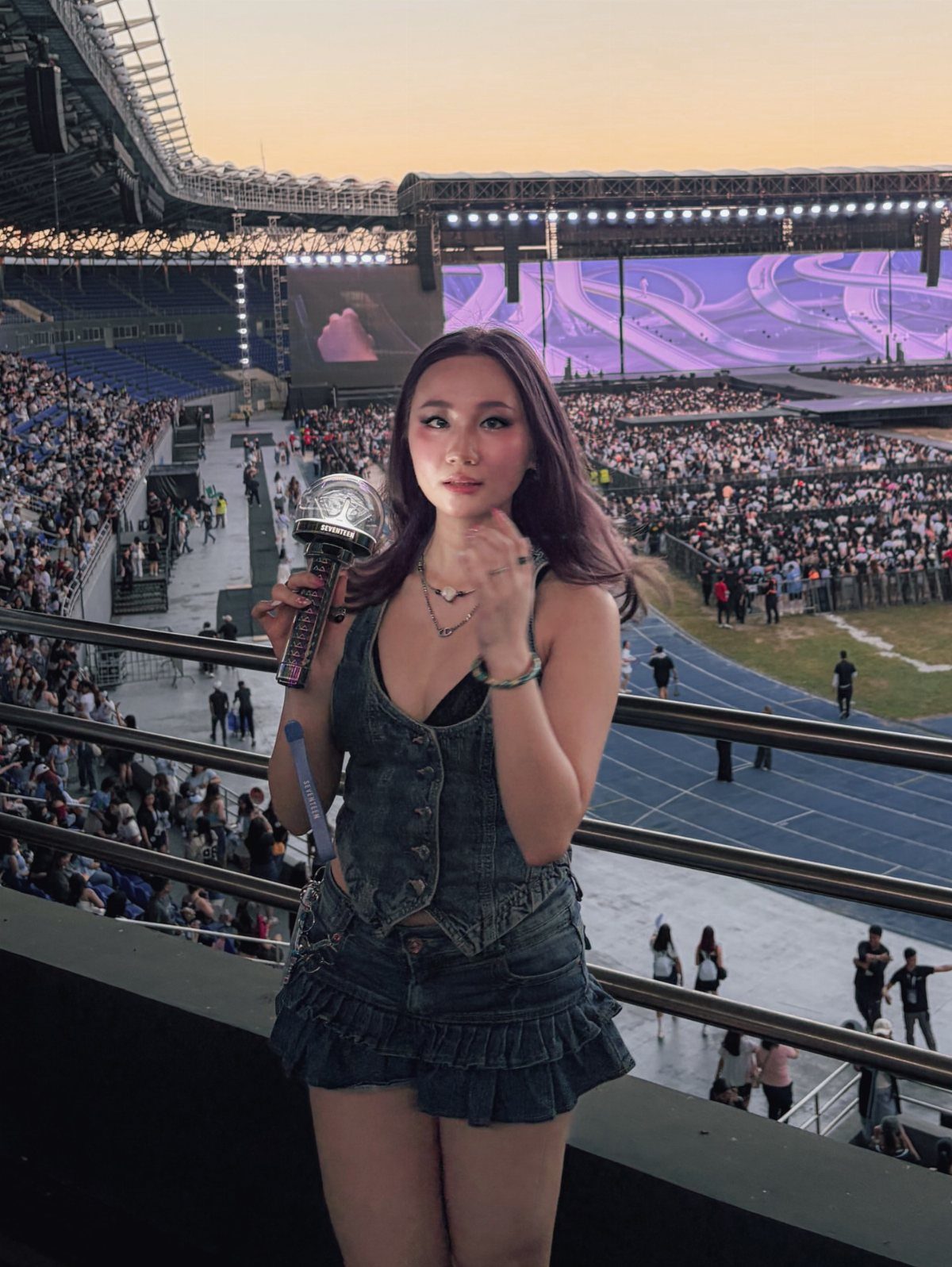12 Seventeen Concert Outfit Ideas for Women to Stand Out 11 Woman in denim vest and ruffled skirt posing at a crowded stadium concert.