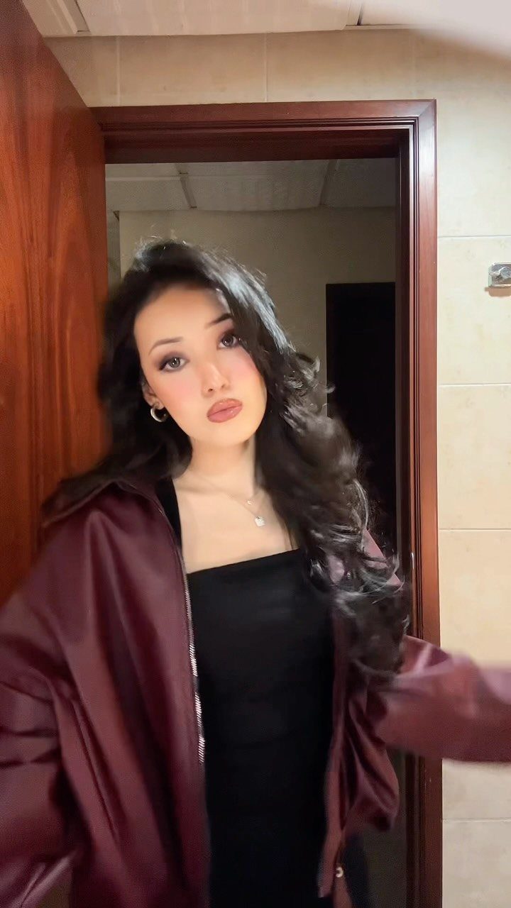 12 Seventeen Concert Outfit Ideas for Women to Stand Out 8 Woman wearing a maroon slouchy bomber jacket over a black top in a doorway.