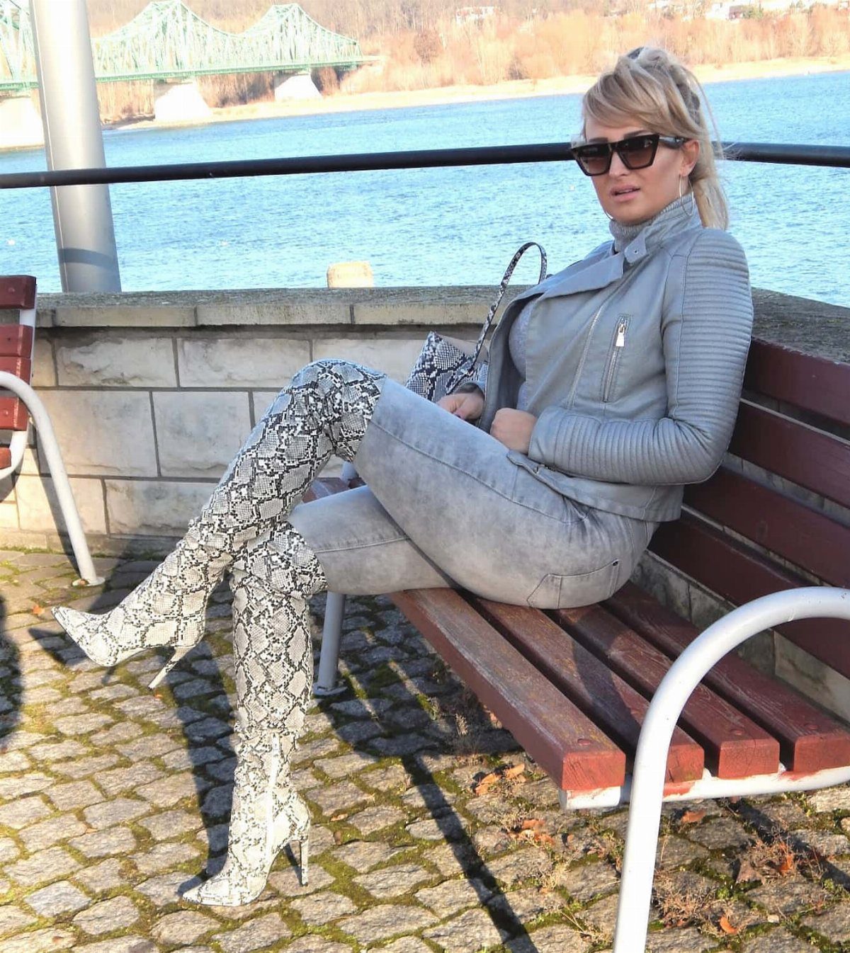 Woman in grey jeans and snakeskin over-the-knee boots sitting on a riverside bench
