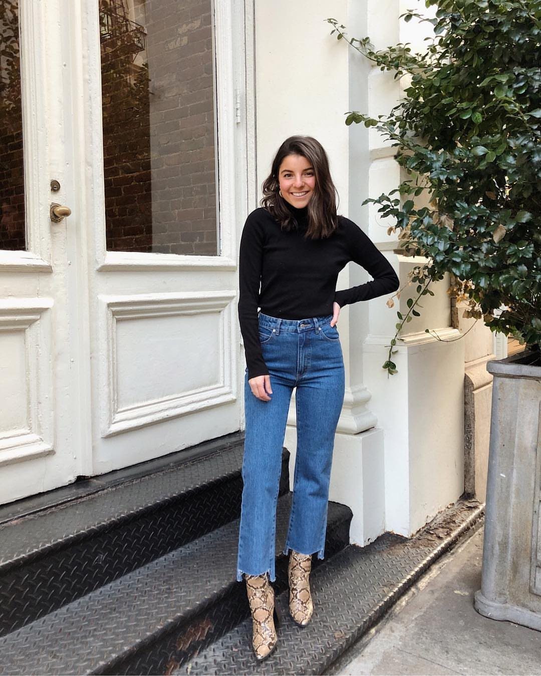 Woman wearing blue straight-leg jeans and tan snakeskin ankle boots standing by a door.