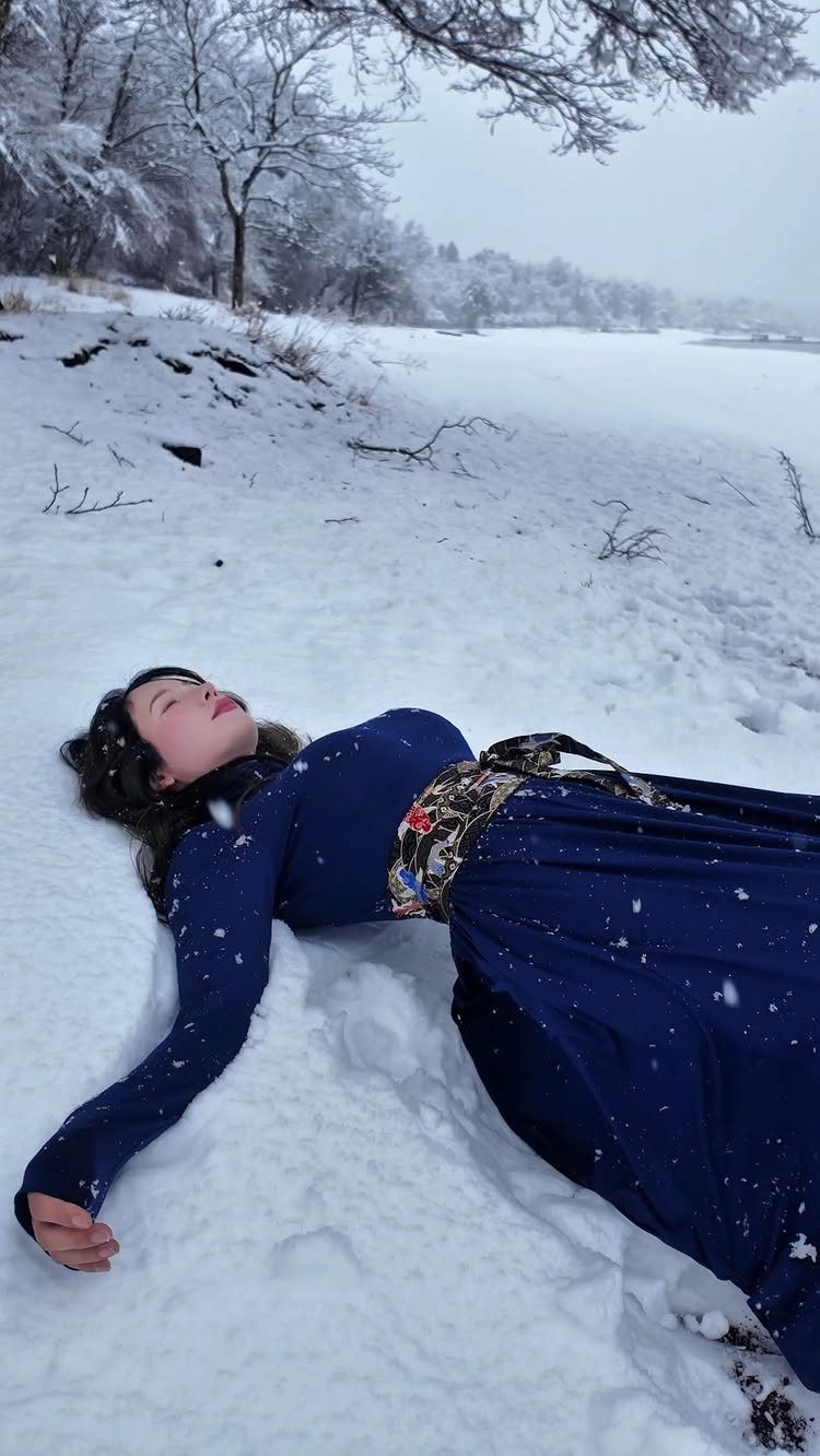 Woman in a royal blue dress lying in deep white snow among winter trees