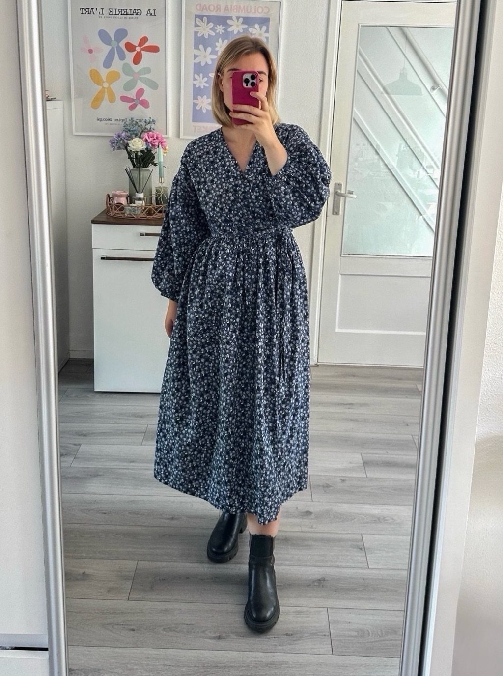 Woman in a navy blue floral midi dress and black boots taking a mirror selfie.