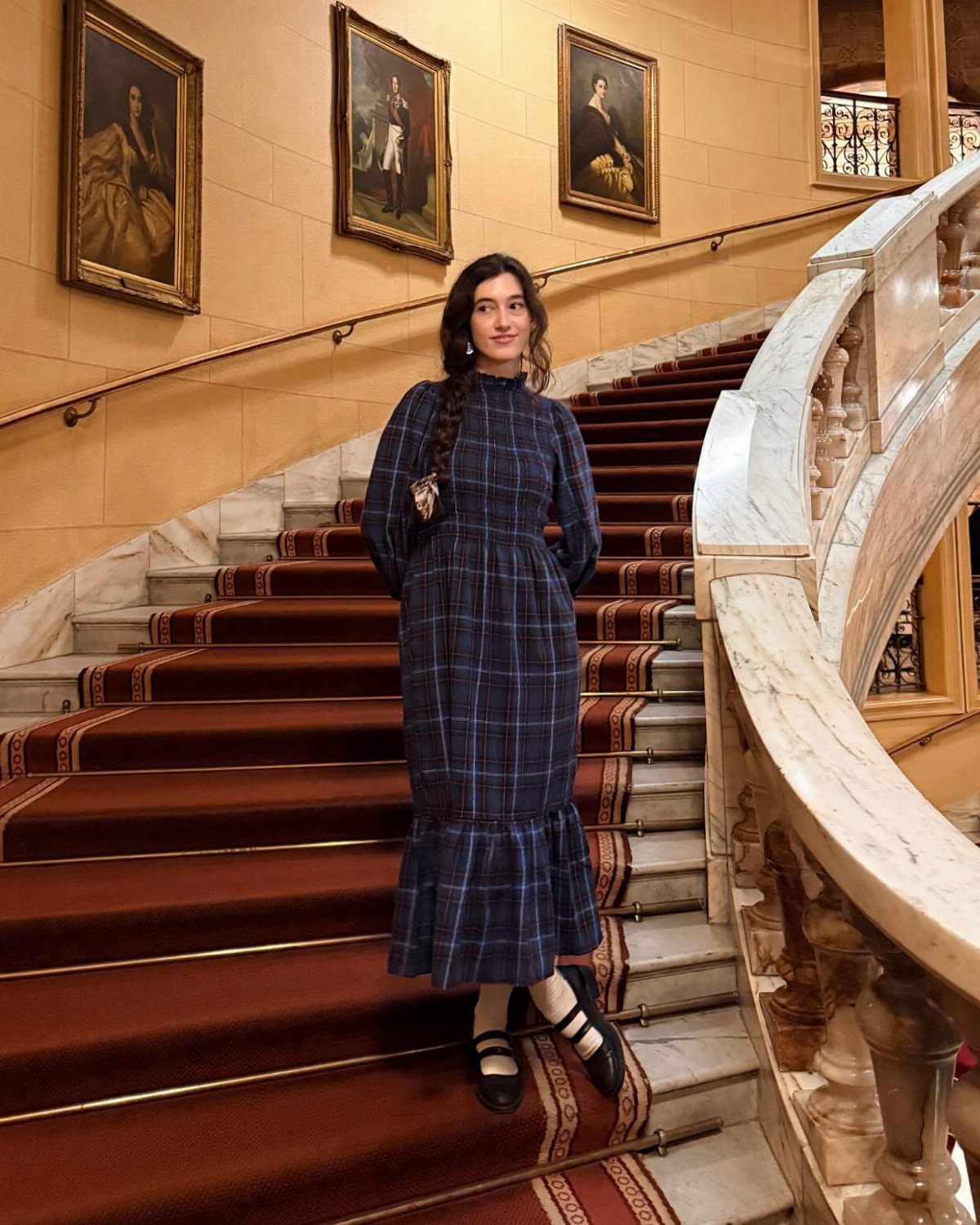 Woman in a navy blue checked long-sleeved dress standing on a grand staircase.
