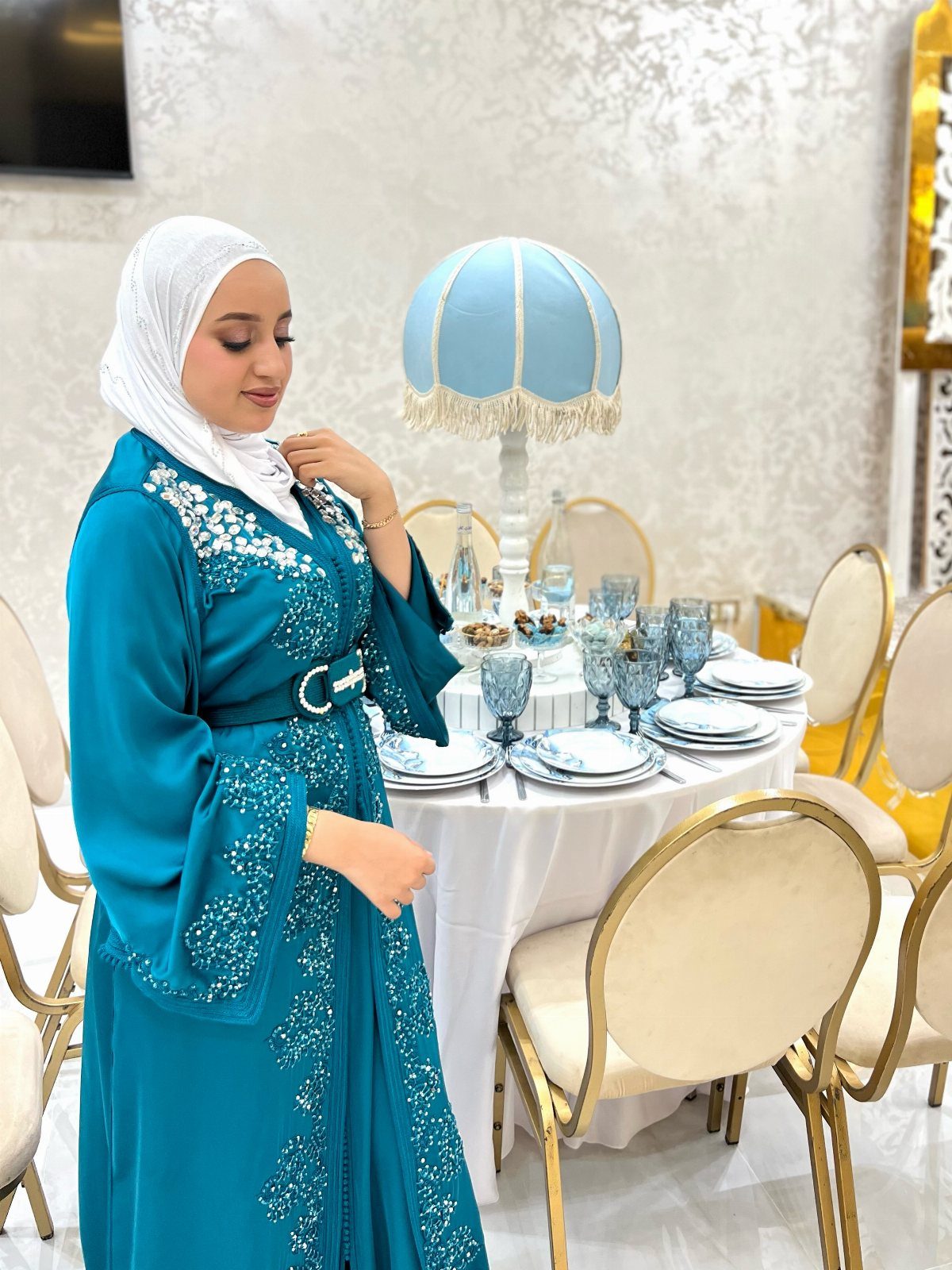 Woman in a teal blue embroidered kaftan dress and white hijab by a table.