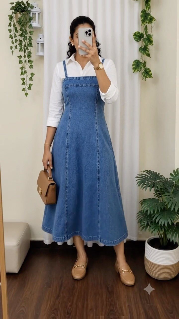 Woman wearing a long blue denim pinafore dress over a white collared shirt.
