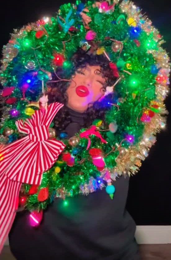 Person wearing a large decorated Christmas wreath as an ugly holiday sweater.